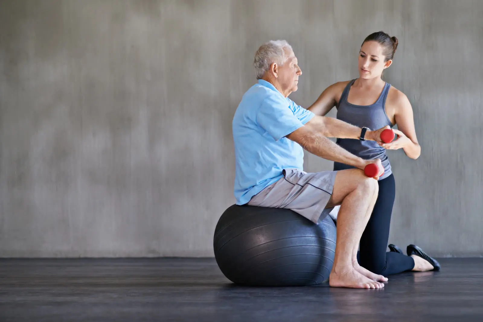 Physiotherapeutin übt mit einem Senior auf dem Gymnastikball mit kleinen Hanteln